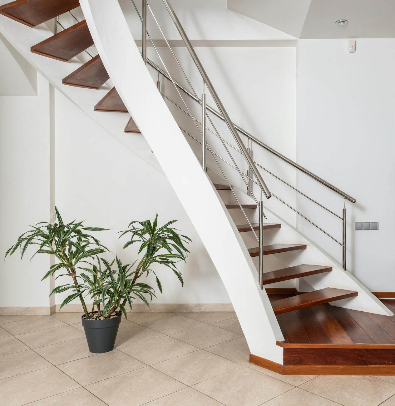 Contemporary wooden staircase with metal railings above ceramic floor with green plant in pot in light house